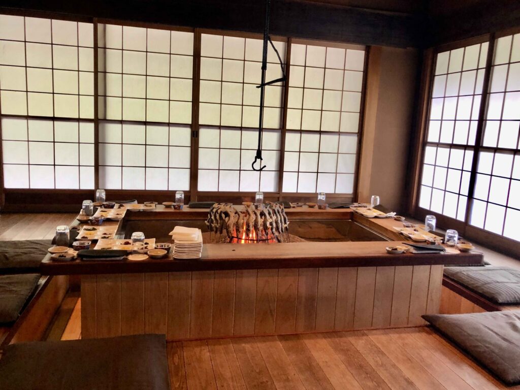 Traditional irori hearth dining room at the famous farmhouse restaurant Yanagiya in Gifu, Japan — featuring skewered ayu fish grilling over charcoal, tatami seating, and shoji windows that frame a serene mountain setting.