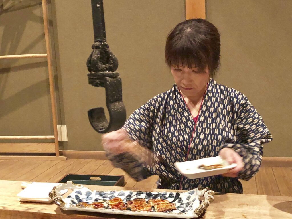 The hostess at the famous Yanagiya farmhouse restaurant in Gifu, Japan, elegantly plating grilled ayu river fish beside the traditional irori hearth — a refined example of Japanese hospitality and mountain cuisine artistry.
