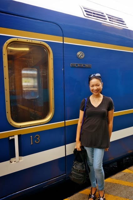 A woman standing beside the iconic deep-blue exterior of The Blue Train at the station, next to a gold-trimmed carriage door marked with the number 13. The polished blue and gold detailing highlights the luxury and heritage of South Africa’s famous rail journey.