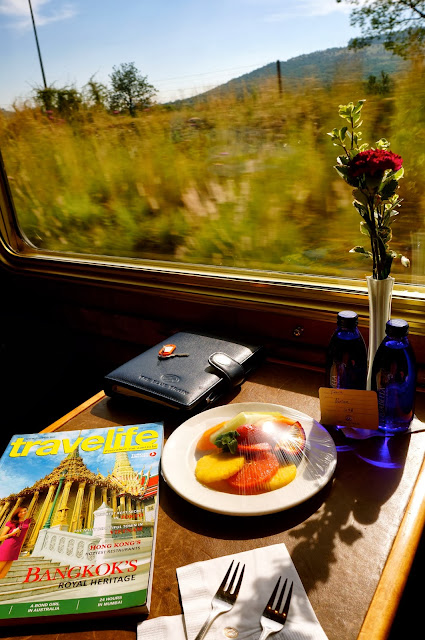 A breakfast setting inside The Blue Train with a plate of fresh fruit, two forks, a Travelife magazine, and a single red rose in a vase placed by the window. Outside, the South African countryside passes in soft, golden light, highlighting the relaxed luxury of the rail journey from Pretoria to Cape Town.