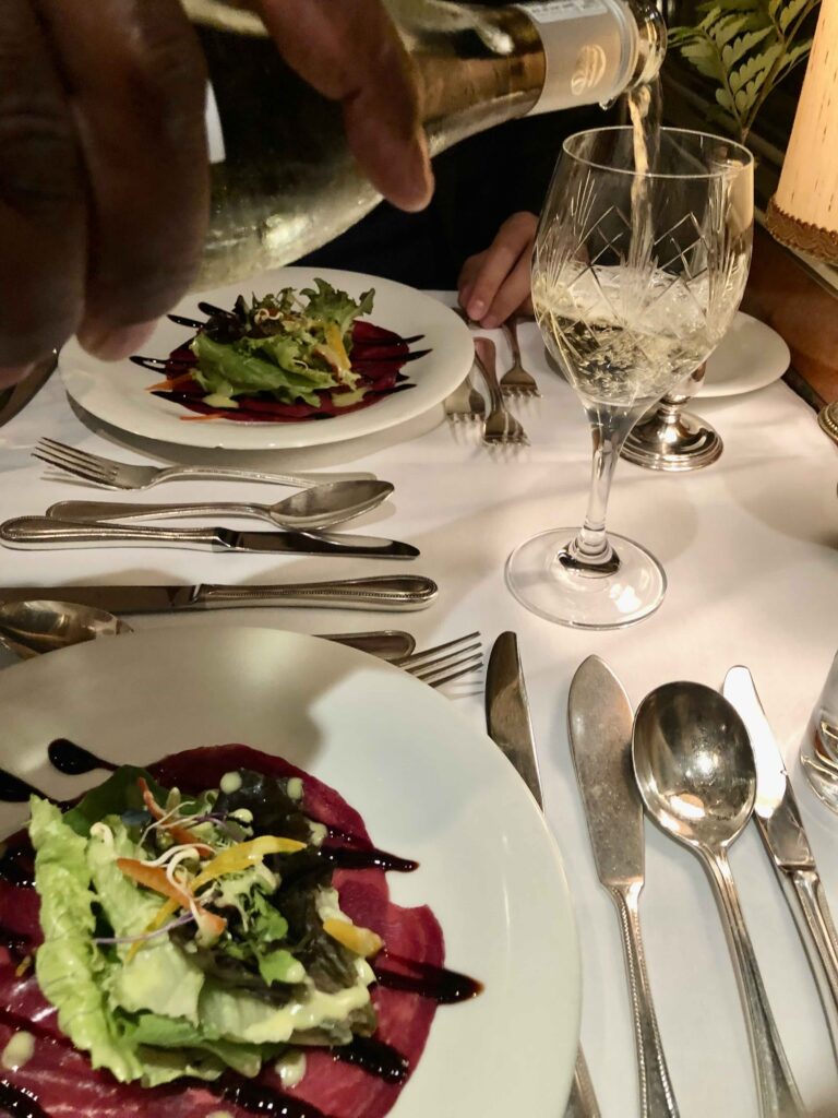 A waiter pouring white wine into a cut-crystal glass beside a plated smoked salmon and salad appetizer in The Blue Train’s elegant dining car, set with polished silverware on a crisp white tablecloth.
