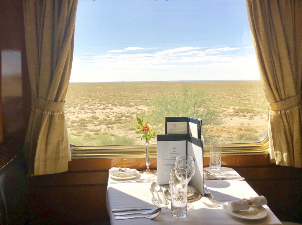 A white table set for lunch onboard The Blue Train, with crystal glasses, silverware, and a menu arranged beside a large window overlooking the wide, sunlit plains of the Northern Cape in South Africa.

