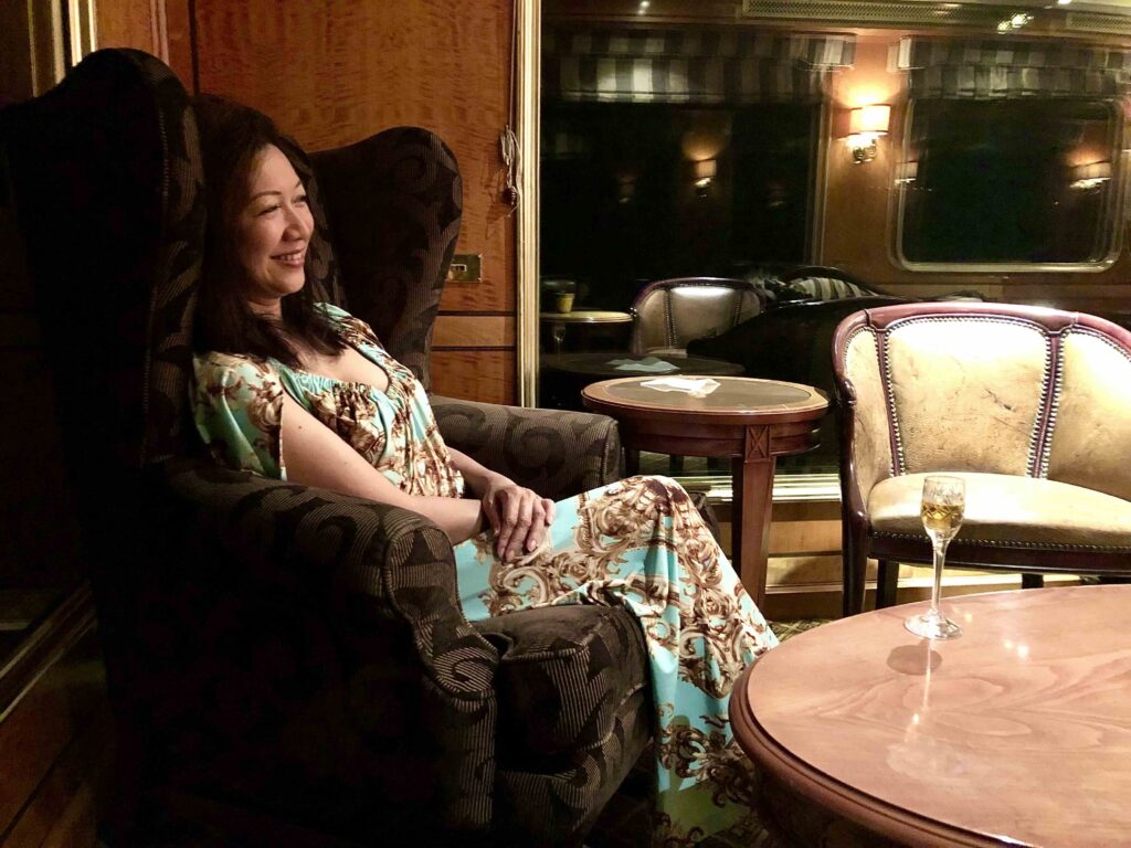 A woman sits elegantly in the lounge car of The Blue Train, surrounded by warm wooden interiors, vintage armchairs, and soft lighting, with a glass of sparkling wine on the table—capturing the relaxed, luxurious atmosphere of South Africa’s iconic rail journey.