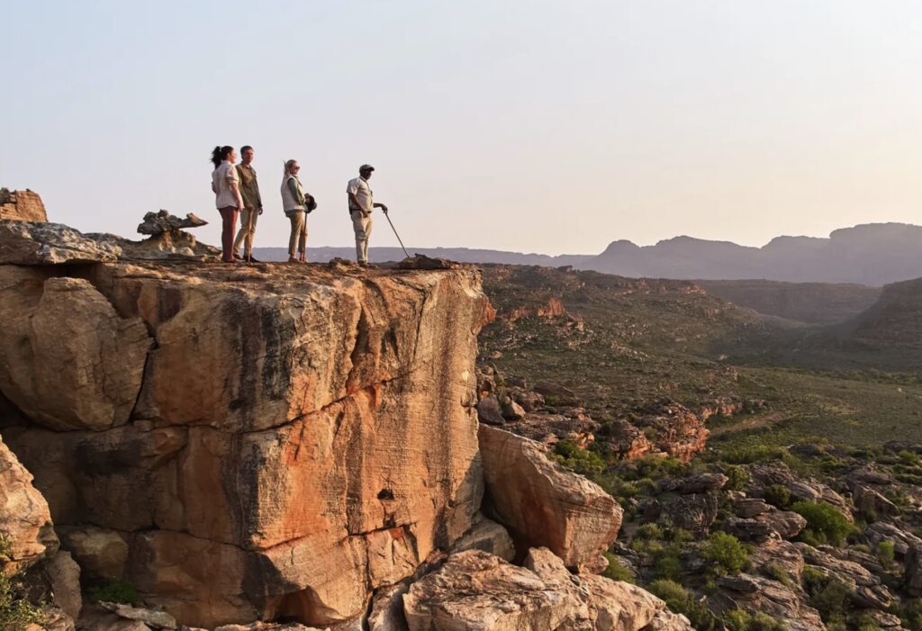 Travelers standing at the edge of a rocky cliff in Bushman’s Kloof, overlooking the vast Cederberg wilderness at sunrise. The scene captures the adventure and serenity of exploring South Africa’s dramatic landscapes within a private nature reserve.