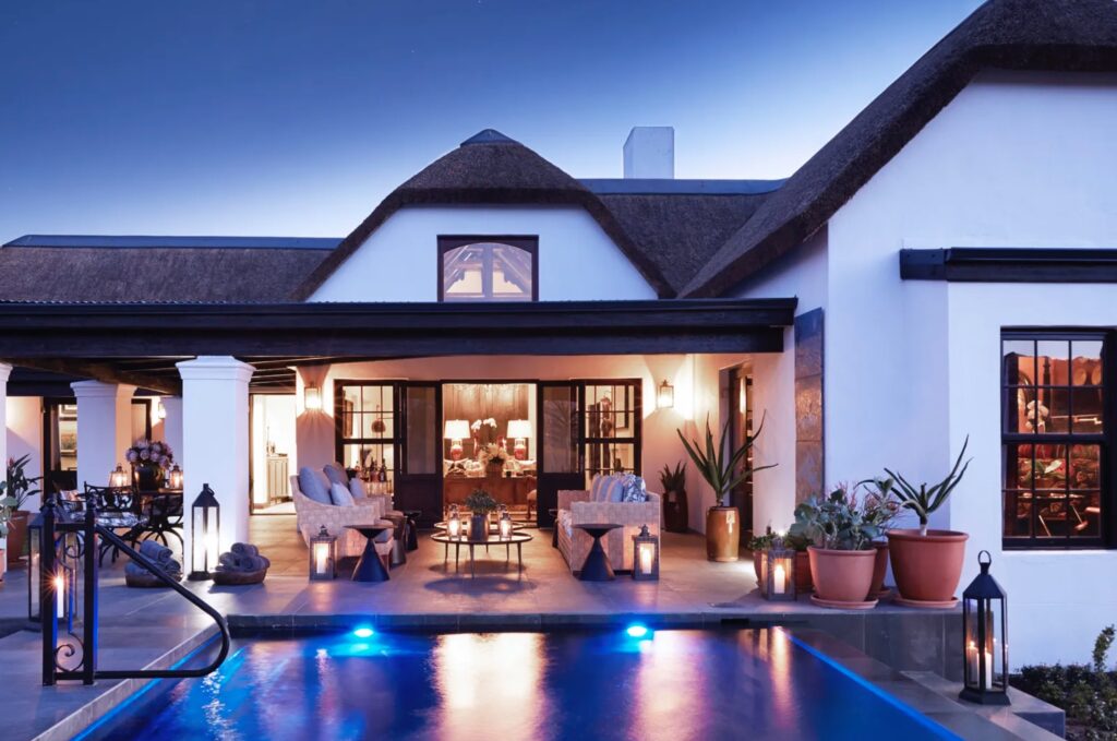 Elegant thatched lodge at Bushman’s Kloof glowing at dusk, with lanterns, pool reflections, and soft golden light spilling from the veranda. The scene captures the serene luxury of South Africa’s Cederberg wilderness — where nature, heritage, and refined hospitality meet under the African twilight.