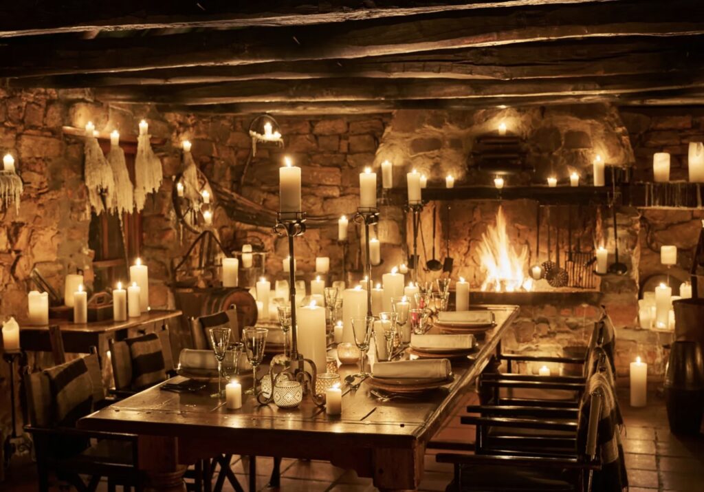 Candlelit dining table inside a rustic stone lodge at Bushman’s Kloof, surrounded by glowing candles and a roaring fireplace. The warm, intimate atmosphere reflects the spirit of South Africa’s Cederberg wilderness — a secluded sanctuary where heritage, nature, and fine dining converge beneath the flicker of firelight. Photo from Bushman's Kloof.