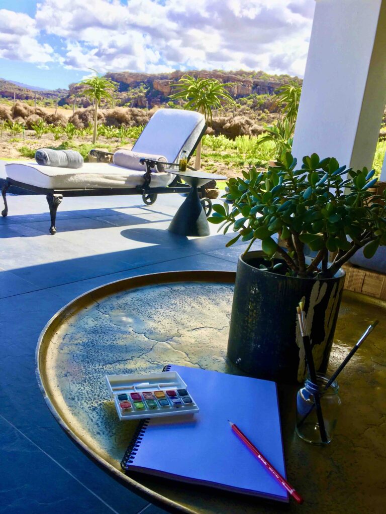 Peaceful terrace scene at Bushman’s Kloof in South Africa’s Cederberg Mountains, featuring a sunlit lounge chair, potted succulents, and a sketchbook on a golden tray. The tranquil outdoor setting invites creativity and reflection amid wilderness views.