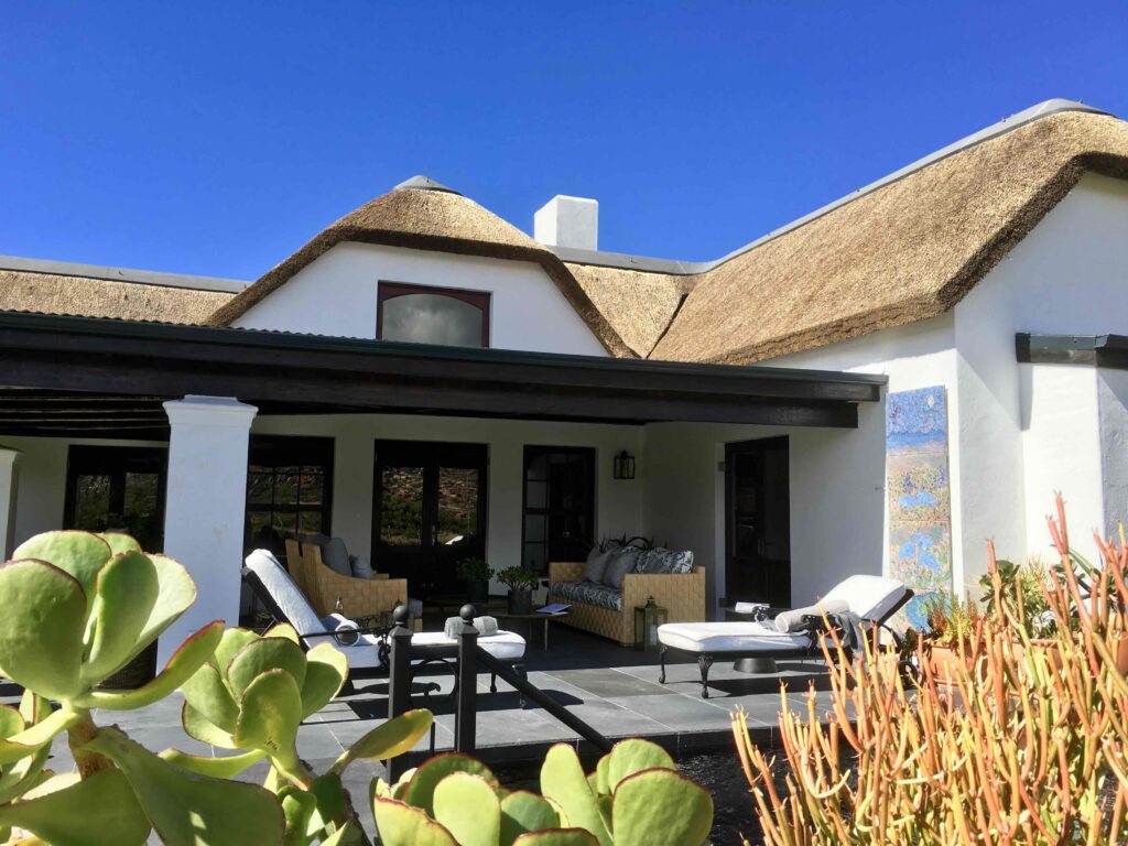 Cape Dutch–style villa at Bushman’s Kloof in South Africa’s Cederberg Mountains, featuring a thatched roof, shaded veranda, and elegant outdoor lounge surrounded by native fynbos. The serene setting reflects the harmony of heritage architecture and wilderness luxury.