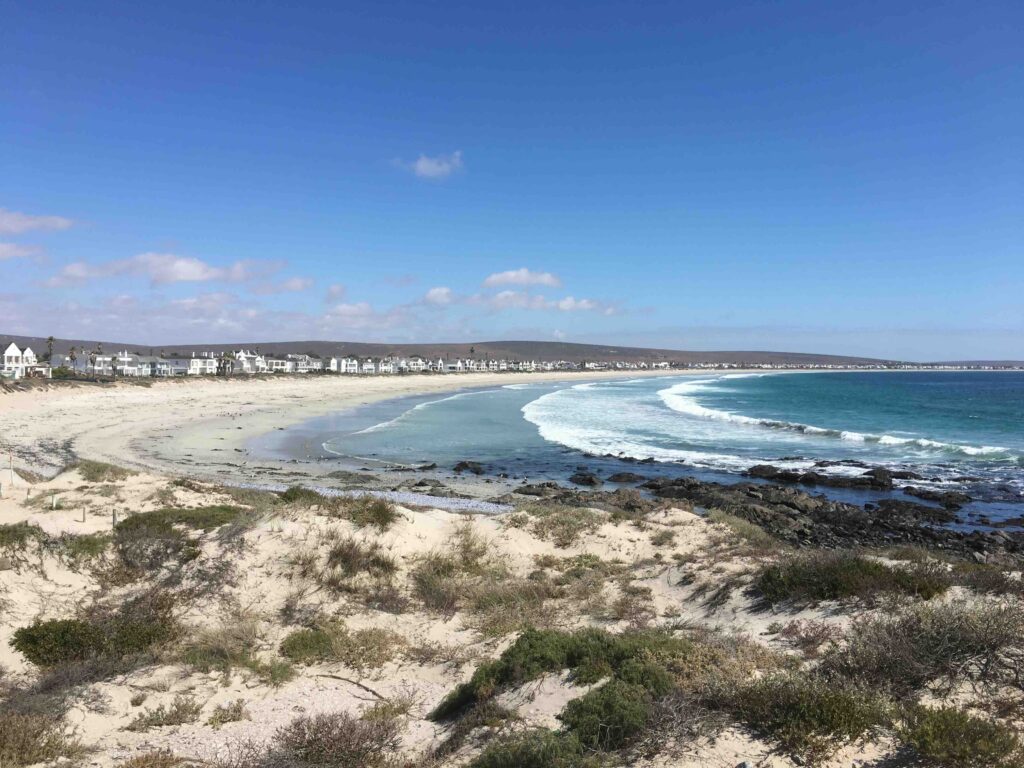 Panoramic view of Shelley Point on South Africa’s West Coast, with white sand dunes, turquoise waves, and coastal homes along the shoreline under a bright blue sky. The tranquil peninsula embodies the wild beauty and serenity of the Western Cape.