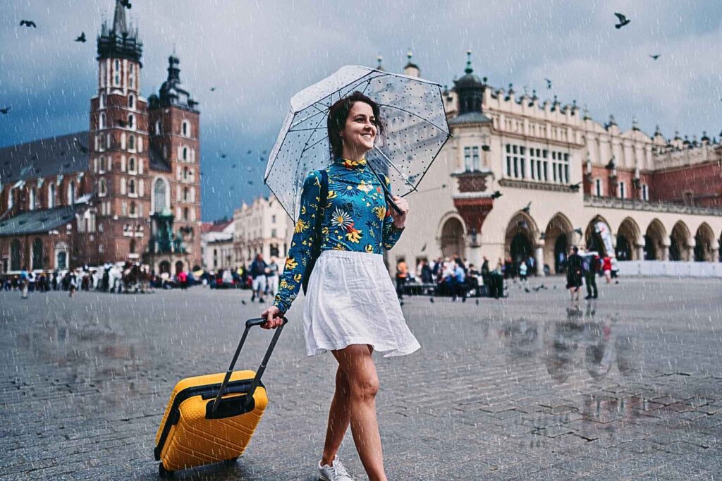 Confident solo female traveler walking through a historic European square under the rain with a yellow suitcase and clear umbrella — representing empowered, fearless, and stylish women’s travel and self-assured exploration abroad.
