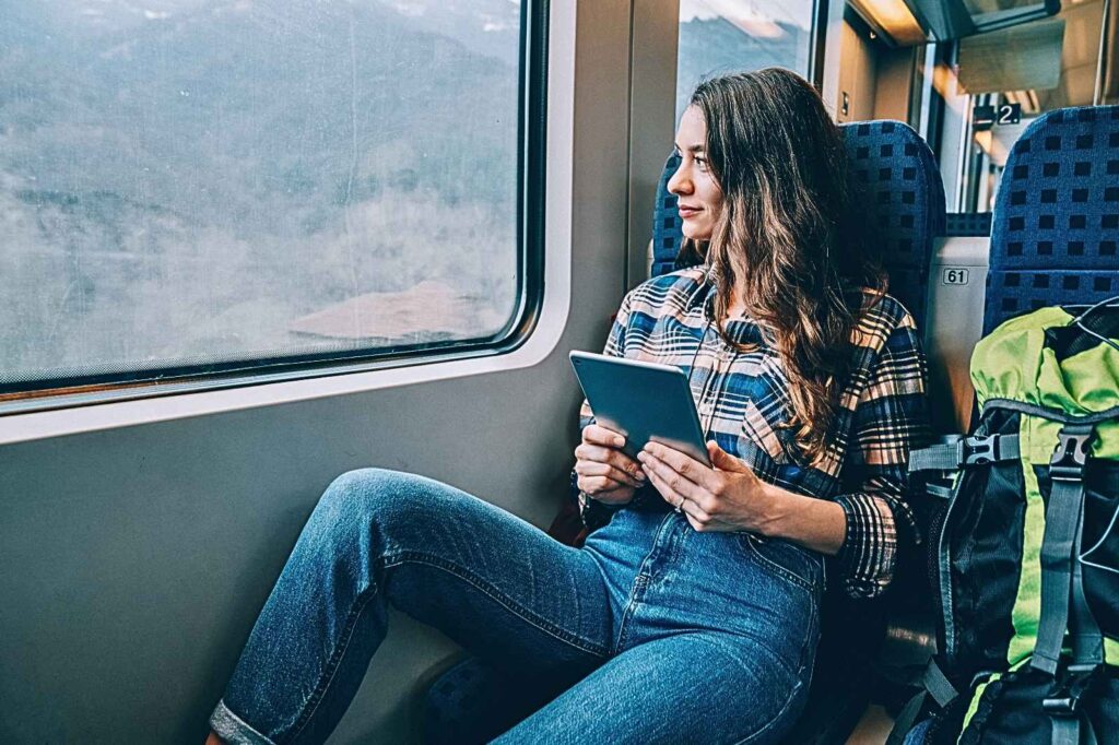 Solo female traveler sitting by a train window with a digital tablet and backpack, enjoying the journey while staying safe and connected — symbol of smart, private, and tech-savvy women’s travel.