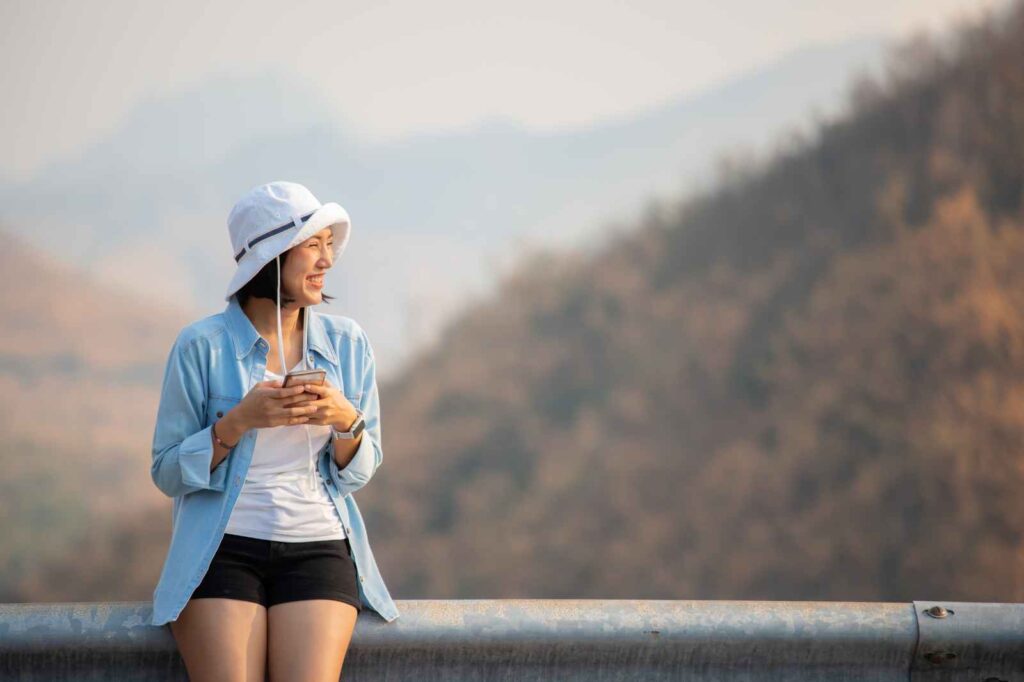 Smiling solo female traveler sitting on a mountain viewpoint with earphones and coffee cup, enjoying peaceful connection and freedom — symbolizing mindful, independent women’s travel and the joy of companionship on one’s own terms.