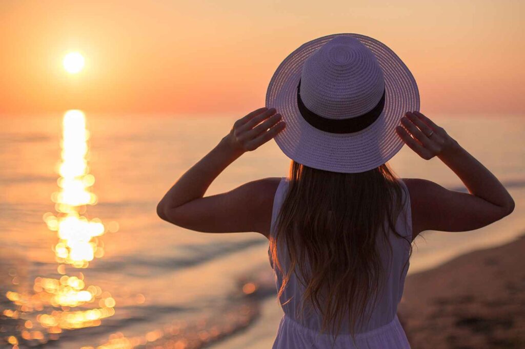 Solo female traveler watching the sunset over the ocean, holding her sun hat with both hands — representing intuition, self-trust, and the calm confidence of independent women’s travel and mindful exploration.