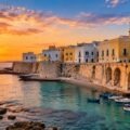 Gallipoli Old Town in Puglia, Italy, with historic waterfront buildings, stone sea walls and fishing boats on the Ionian Sea at sunset.