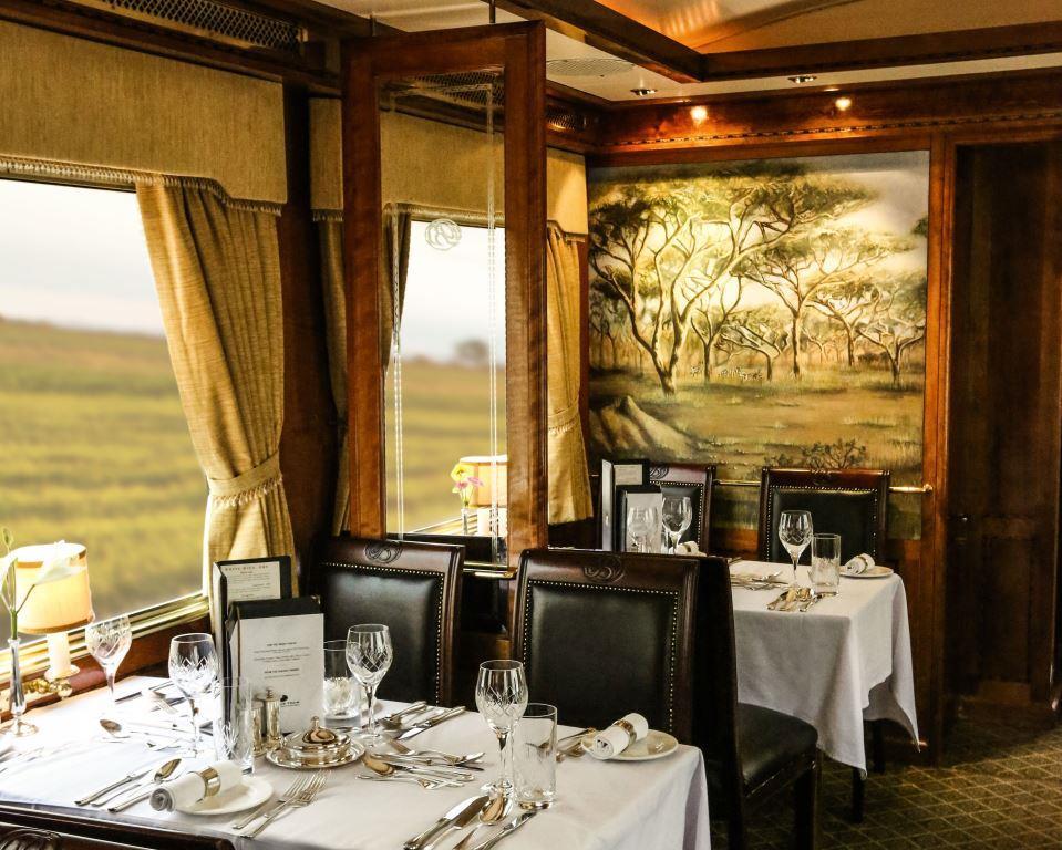 A lunch table set with crystal glasses, polished silverware, and a standing menu inside The Blue Train, framed by gold curtains and a large window overlooking South Africa’s rugged mountains and open countryside.