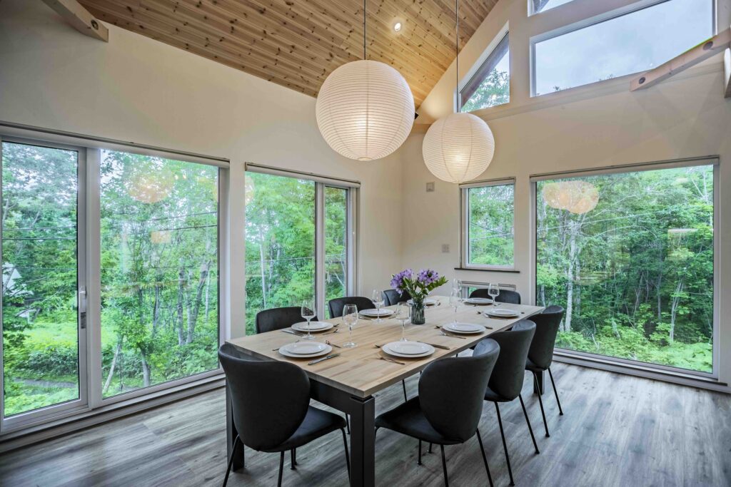 Dining area at Soga Forest Chalet in Niseko, Hokkaido, with a wooden dining table, pendant lights and large windows overlooking the forest.