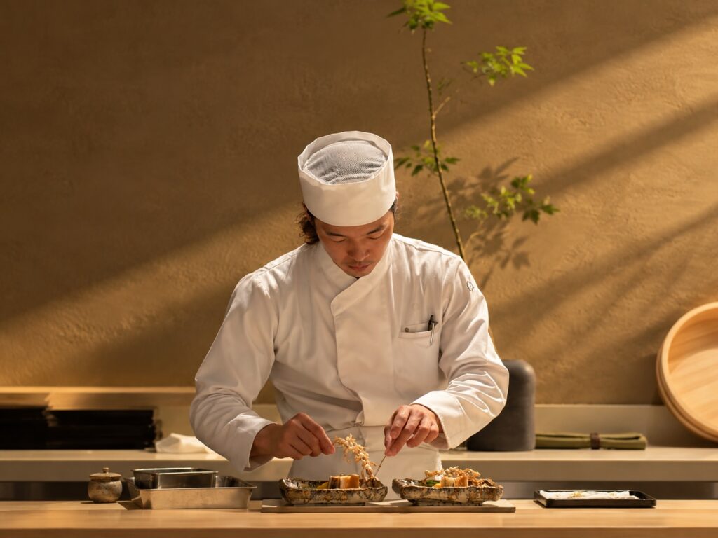Japanese chef in a white uniform and traditional chef's hat preparing food at a restaurant counter at THE PASONA Natureverse Retreat on Awaji Island.