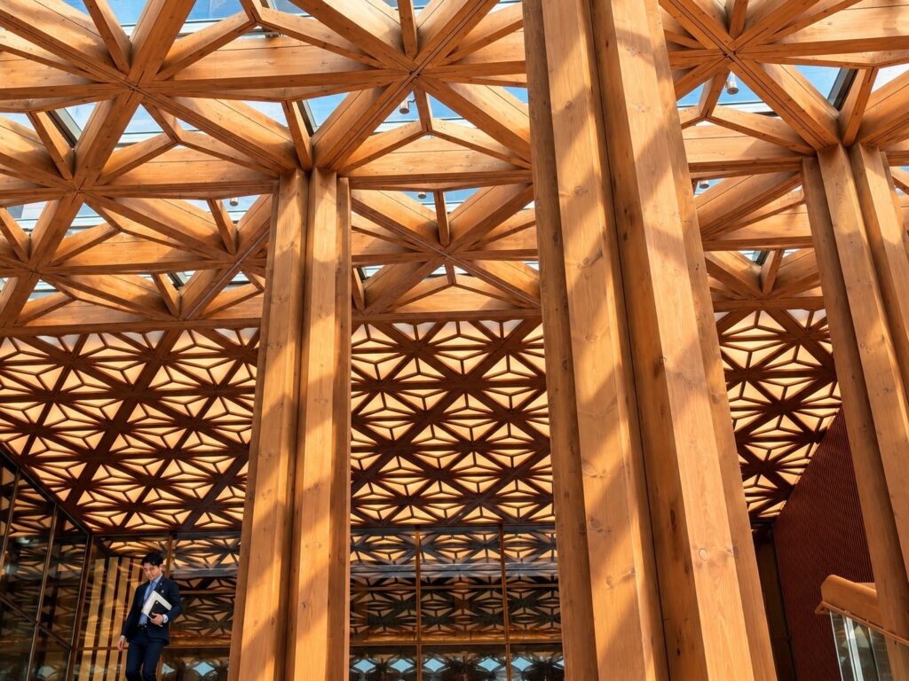 Intricate geometric timber lattice ceiling and towering wooden columns at THE PASONA natureverse retreat on Awaji Island, Japan, designed by architect Shigeru Ban.